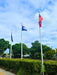 Three flagpoles outdoors with flags flying above a trimmed green hedge under a blue sky with white clouds. One flag appears to be the Queensland state flag, and another visible flag has a red and yellow design.