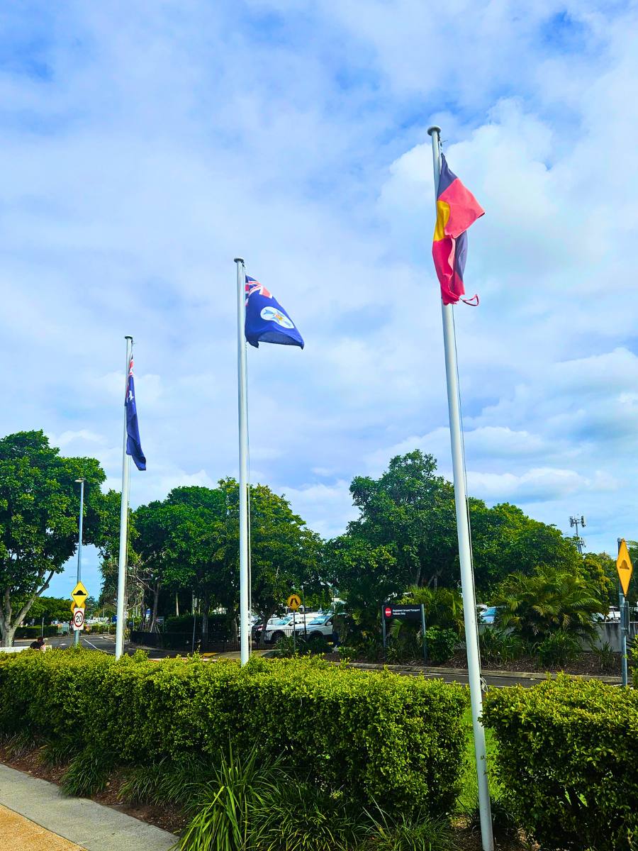 Three flagpoles outdoors with flags flying above a trimmed green hedge under a blue sky with white clouds. One flag appears to be the Queensland state flag, and another visible flag has a red and yellow design.