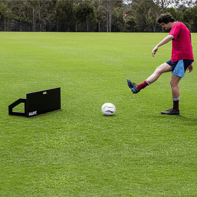 Man kicking ball into HART Soccer Rebound Board whilst on soccer field on green grass