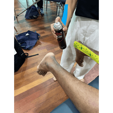 Strapit Xtra Tac Adhesive Spray – person applying spray to another person’s leg on wooden floor in sports facility.